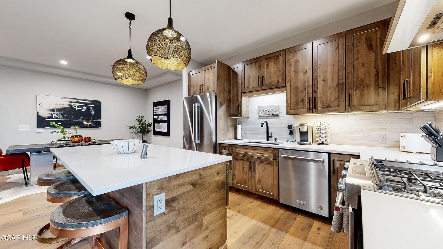 This is a well-lit kitchen featuring wooden cabinetry and stainless steel appliances. A large island with a white countertop and bar stools anchors the space, while pendant lights hang above. The kitchen also includes a stainless steel refrigerator, dishwasher, and a gas range.