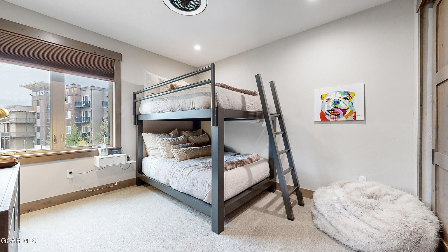 This is a cozy guest bedroom featuring a bunk bed with neutral-toned bedding and decorative pillows. A colorful dog portrait adds a playful touch to the room, while a large, fluffy beanbag chair offers a comfortable seating option. The room is well-lit and has a warm, inviting atmosphere.