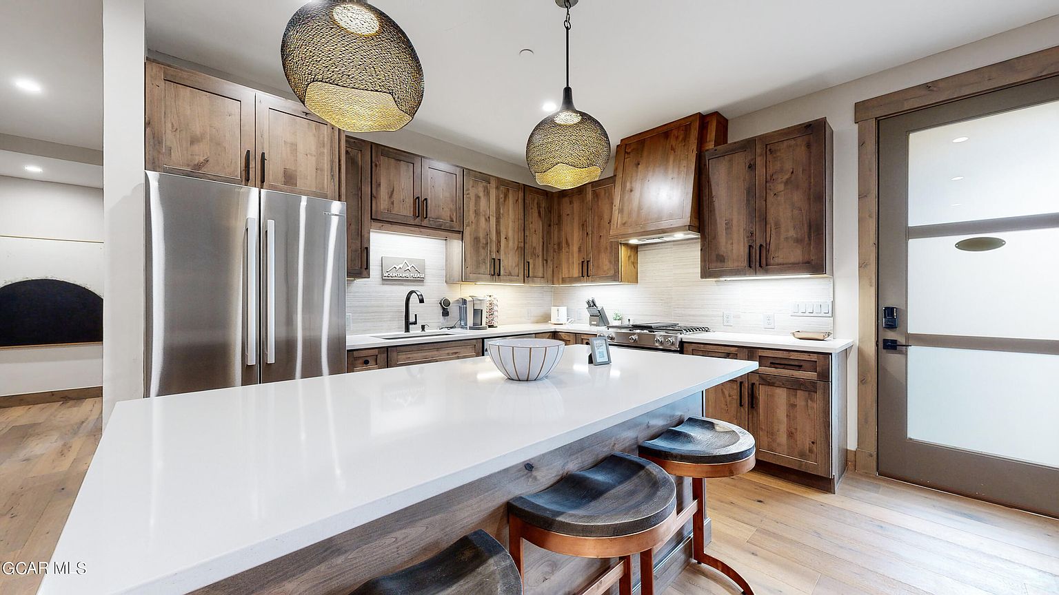 This is a well-lit kitchen featuring wooden cabinetry, stainless steel appliances, and a large white countertop island with bar stool seating. The kitchen has a modern rustic style with unique pendant lighting and a clean, organized appearance. The perspective is from a wide angle, showcasing the entire kitchen space.
