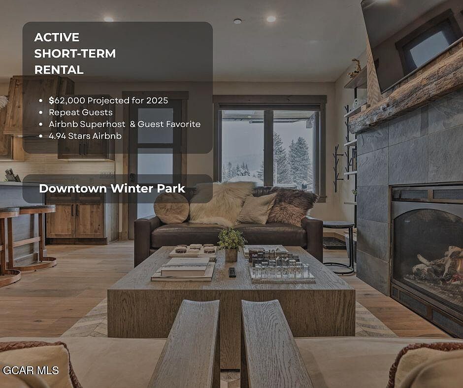 This is an interior shot of a cozy living room, likely in a short-term rental property. The room features a stone fireplace, a comfortable leather sofa with decorative pillows, and a wooden coffee table. Large windows offer a view of a snowy landscape, enhancing the room's inviting and relaxing atmosphere.