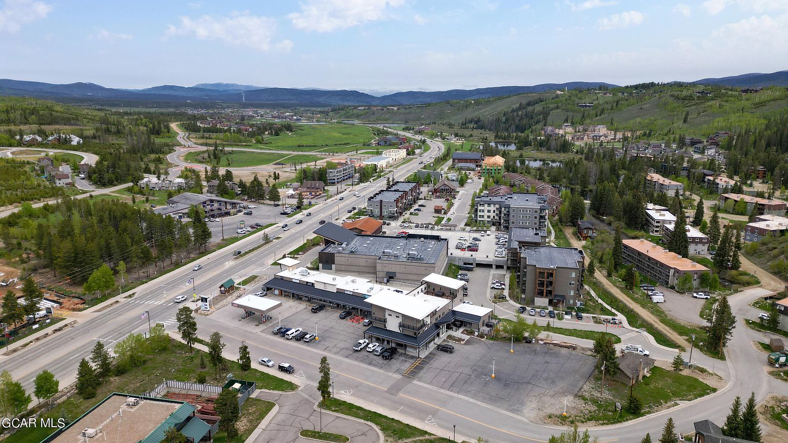 This aerial view showcases a commercial area with a mix of buildings, parking lots, and surrounding natural landscape. The buildings feature modern architectural styles with flat roofs and a combination of light and dark exterior finishes. The scene is set against a backdrop of rolling hills and mountains, providing a sense of location and context.