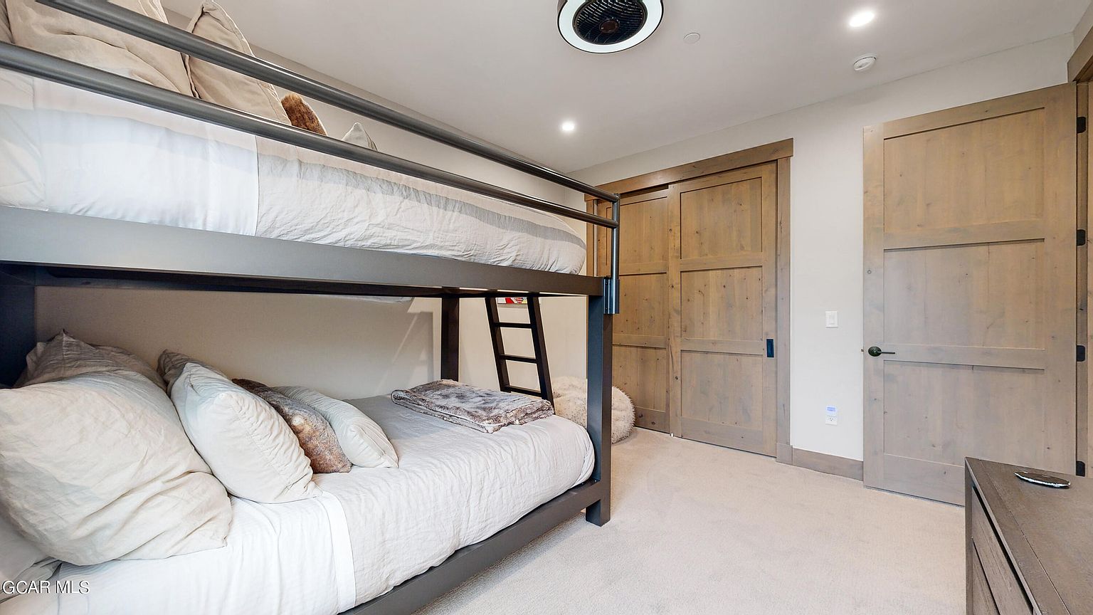 This is a cozy guest bedroom featuring a bunk bed with a gray metal frame and neutral bedding. The room has light-colored walls, carpeted flooring, and wooden doors, creating a warm and inviting atmosphere. A ceiling fan and recessed lighting provide ample illumination.