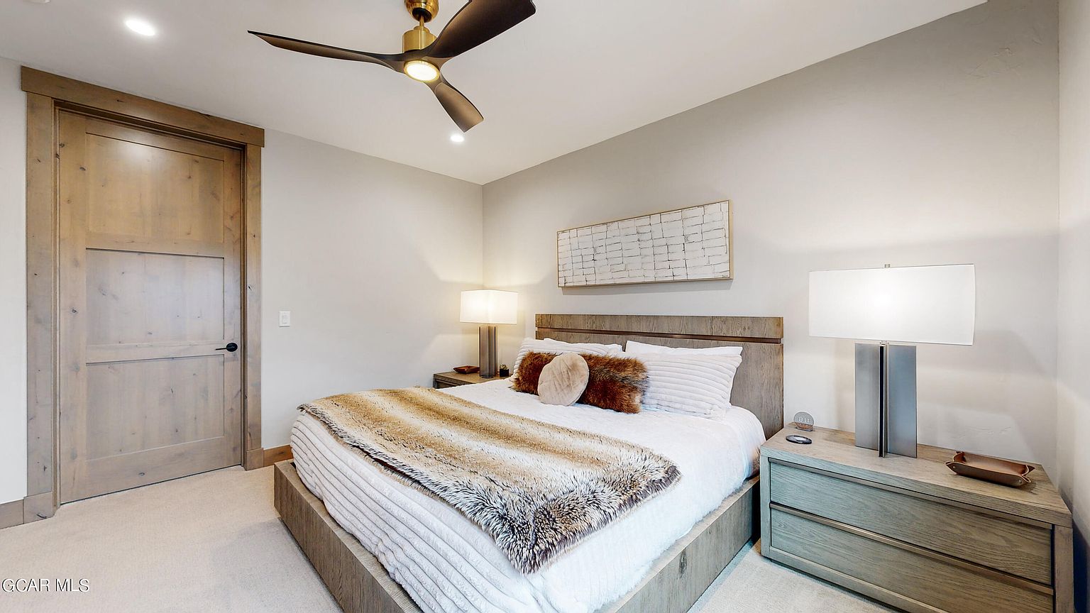 This is a cozy primary bedroom featuring a wooden bed frame with a plush white comforter and a faux fur throw. The room is decorated with neutral tones, complemented by wooden nightstands and lamps. A modern ceiling fan and artwork add to the room's contemporary yet inviting atmosphere.