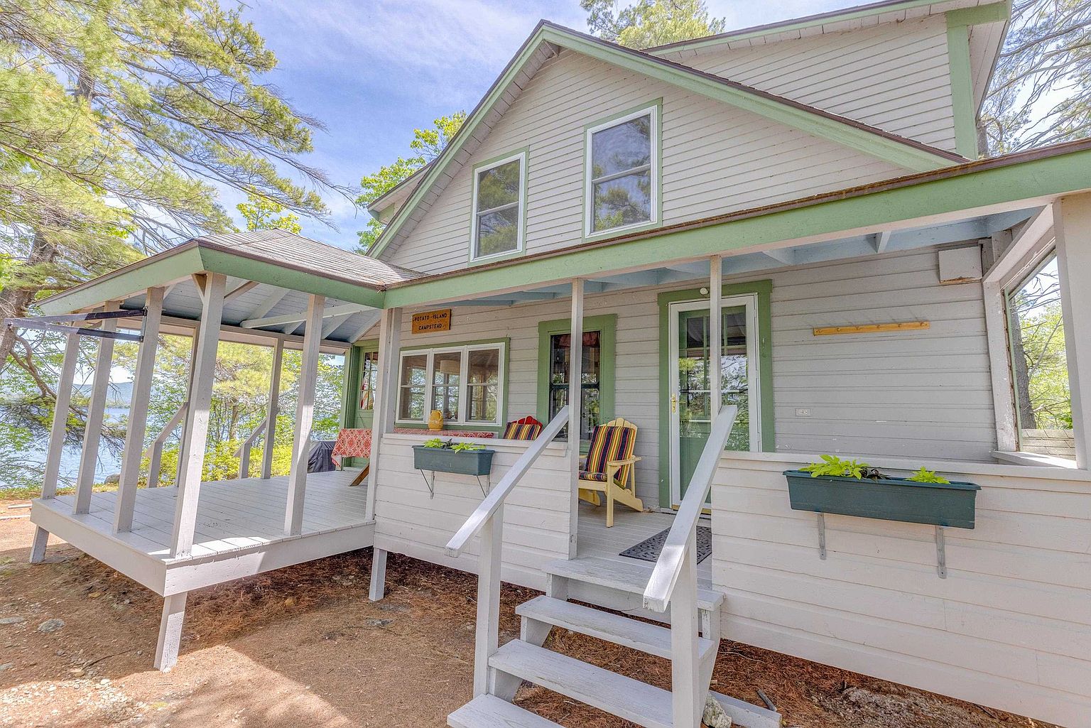 This charming, light-gray wood-sided cottage features a wraparound porch with green trim and a welcoming set of stairs leading to the entrance. The home is nestled in a wooded setting with mature trees and a glimpse of a lake in the background, creating a peaceful, rustic atmosphere. A small sign above the porch identifies the property as 'Potato Island Campstead,' highlighting its unique, secluded character.