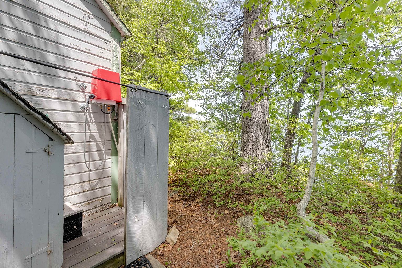 This outdoor shower area is attached to the side of a light-colored, wood-sided structure, featuring a bright red water heater unit and a simple shower head. A wooden privacy screen partially encloses the shower space, which sits on a small wooden deck. The setting is rustic and secluded, surrounded by lush green trees and foliage with a glimpse of a lake in the background.