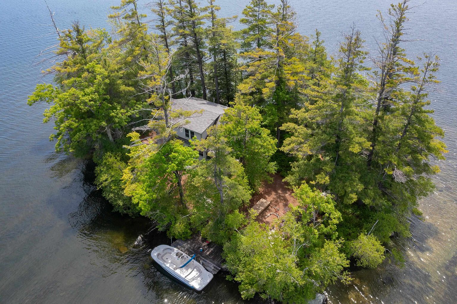 This aerial perspective captures a secluded island property surrounded by calm water, featuring a private house nestled among dense, mature trees. A wooden dock extends from the shoreline, where a small motorboat is moored, providing a clear sense of the property's waterfront lifestyle. The lush greenery and natural landscape create a serene and private atmosphere, highlighting the unique appeal of this island retreat.