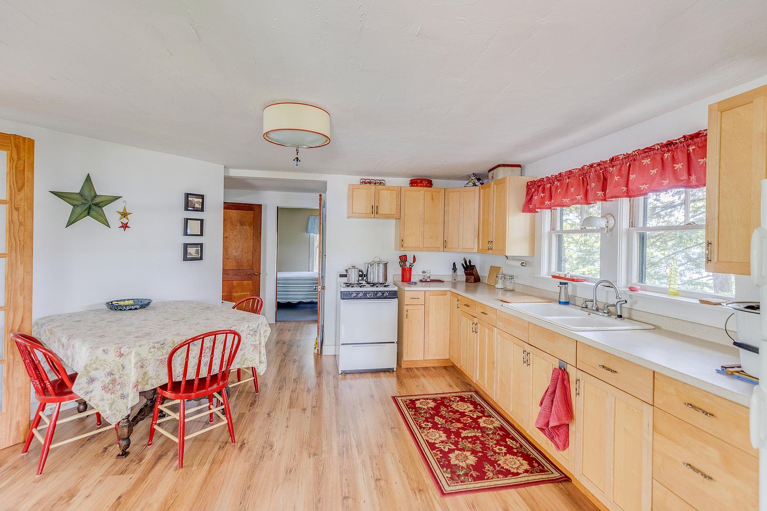 This bright, eat-in kitchen features light wood cabinetry, a white stove, and a double-basin sink positioned beneath windows with red patterned valances. A round dining table with red chairs sits to the left, creating a cozy breakfast nook area. The space is finished with light-toned wood flooring and a decorative red floral rug, offering a warm and traditional cottage-style aesthetic.