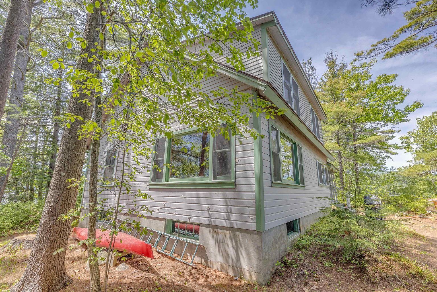 This side view of a two-story home showcases light-colored horizontal siding accented by green trim around the windows and corners. The structure features a concrete foundation, a prominent multi-pane window on the main level, and a red canoe resting on the ground near a ladder. The house is nestled in a wooded setting with mature trees surrounding the property, creating a secluded and natural atmosphere.