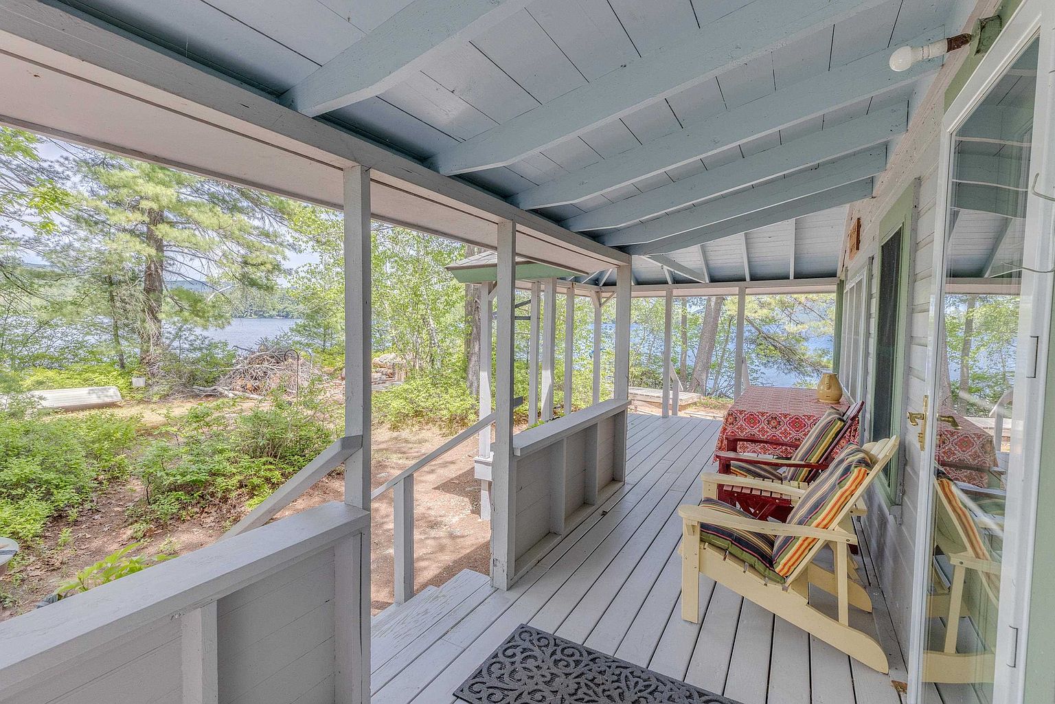 This covered wooden deck offers a serene, elevated view of a lake surrounded by lush forest. The space is furnished with two striped wooden chairs and a table covered in a patterned red cloth, creating a cozy spot for outdoor relaxation. The light blue painted ceiling and open-railing design enhance the airy, rustic atmosphere of this lakeside retreat.