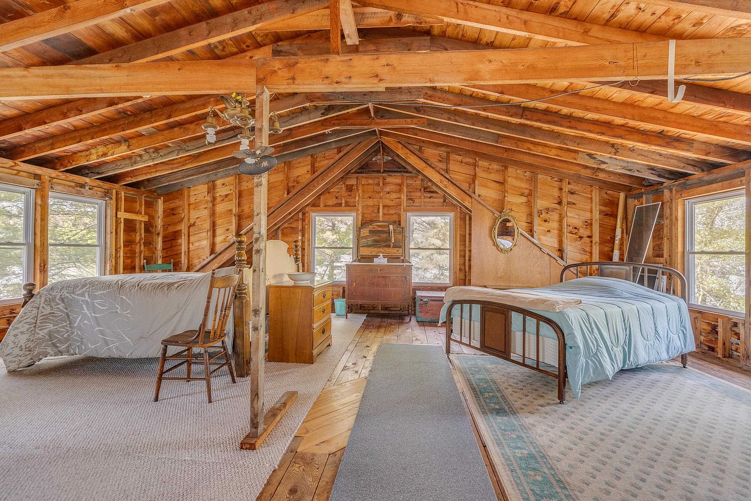 This rustic attic bedroom features exposed wooden rafters and walls, creating a cozy, cabin-like atmosphere. The space is furnished with two beds, a wooden dresser, and a vintage-style vanity, all arranged under the sloping roofline. Multiple windows allow natural light to fill the room, highlighting the warm wood tones and the open, airy layout.