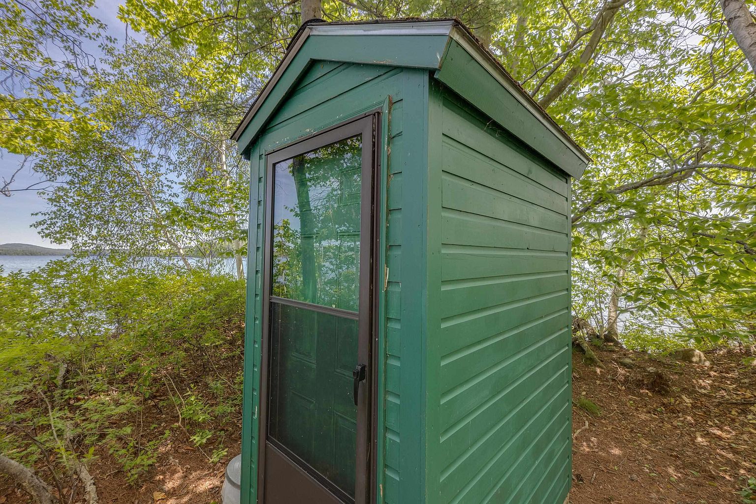 This image captures a small, green-painted wooden outhouse or storage shed nestled in a wooded area near a lake. The structure features a dark-framed screen door and horizontal siding, set against a backdrop of lush green trees and a glimpse of the water. The perspective is a close-up side angle, emphasizing the rustic, secluded nature of the property.