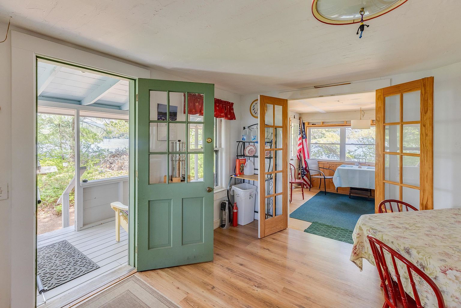 This interior view captures a dining area that flows into a sun-drenched porch and an adjacent room through French doors. The space features light wood-look flooring, a floral-patterned tablecloth on a dining table, and a green-painted door leading to a screened-in outdoor area with lake views. The atmosphere is bright and casual, emphasizing a seamless connection between the indoor living space and the natural surroundings.