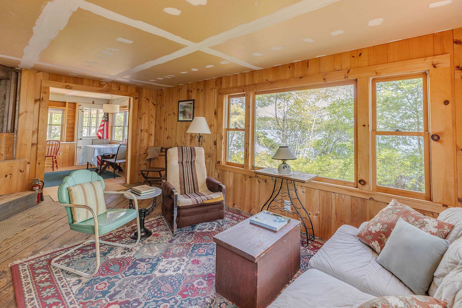This cozy, rustic living room features warm wood-paneled walls and a large, patterned area rug that anchors the seating area. The space includes a light-colored sofa, a vintage-style armchair, and a classic upholstered chair, all arranged around a simple wooden coffee table. Large windows provide a scenic view of the surrounding trees and water, while an open doorway leads to an adjacent dining area with a view of an American flag.