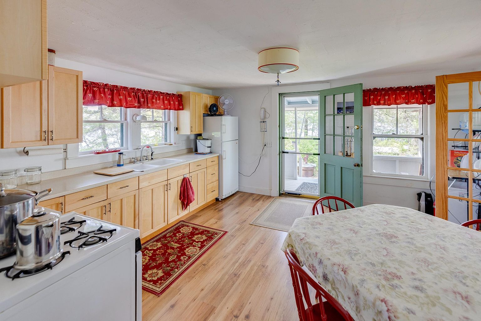 This cozy, rustic-style kitchen features light wood cabinetry, a white stove, and a double-basin sink set against a window with red patterned valances. A dining area with a floral-patterned tablecloth sits in the foreground, while a green-painted door leads out to a screened porch, providing a bright, airy feel. The space is finished with light-toned wood flooring and a red area rug, creating a warm and inviting atmosphere.
