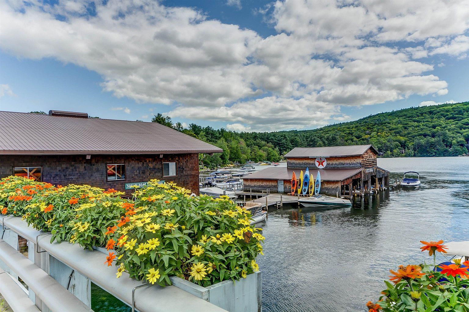 This scenic waterfront view captures a rustic boat house and marina complex nestled against a lush, tree-covered hillside. In the foreground, a vibrant planter box filled with yellow and orange zinnias frames the scene, while a collection of colorful kayaks hangs on the side of the wooden structure. The calm water reflects the blue sky and clouds, creating a peaceful and inviting atmosphere typical of a lakeside recreational property.