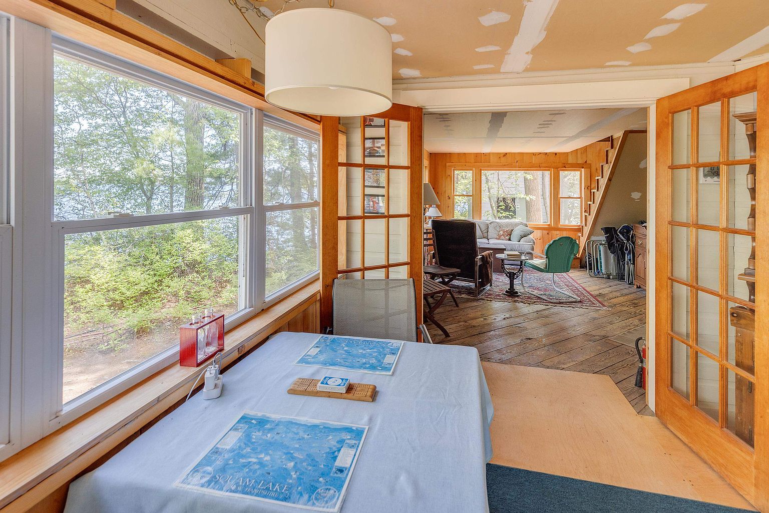 This sunroom features a casual dining area in the foreground with a table set for games, overlooking a scenic lake through large windows. The space transitions into an adjacent living area through glass-paned double doors, showcasing rustic wood-paneled walls and hardwood flooring. The room offers a bright, airy atmosphere with a view of the surrounding forest and water, creating a peaceful retreat.