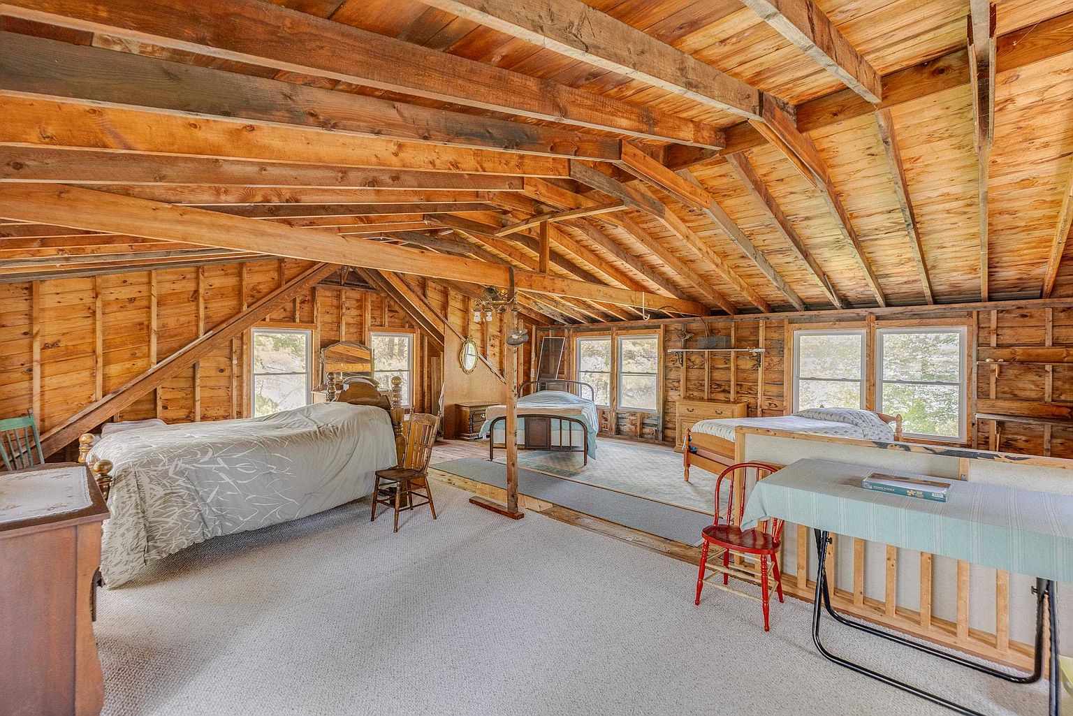 This spacious attic bedroom features exposed wooden rafters and walls, creating a rustic, cabin-like atmosphere. The room is furnished with three beds, a wooden chair, and a folding table, all set against a backdrop of light-colored carpeting. Multiple windows allow natural light to fill the open-concept space, which is divided by a low wooden railing.
