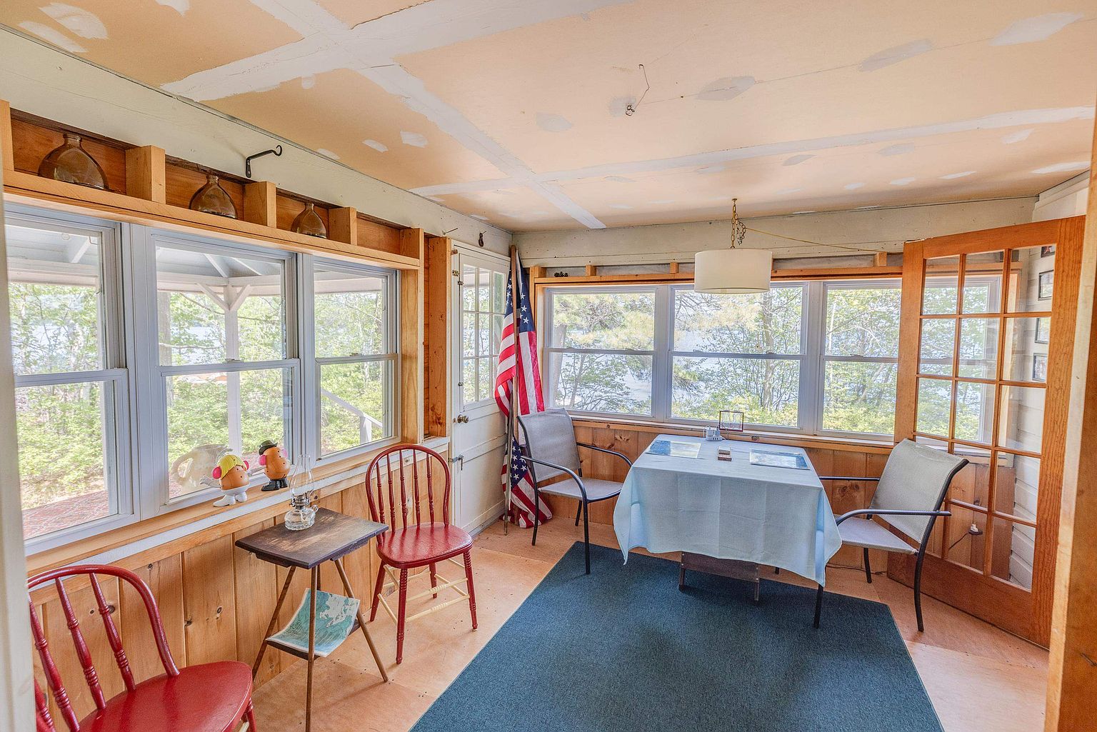 This sunroom features a rustic, unfinished aesthetic with exposed wood-paneled walls and a ceiling currently undergoing renovation. Large windows wrap around the space, providing abundant natural light and scenic views of the surrounding trees and water. The room is furnished with a dining table, several chairs, and a small side table, creating a casual, relaxed atmosphere.