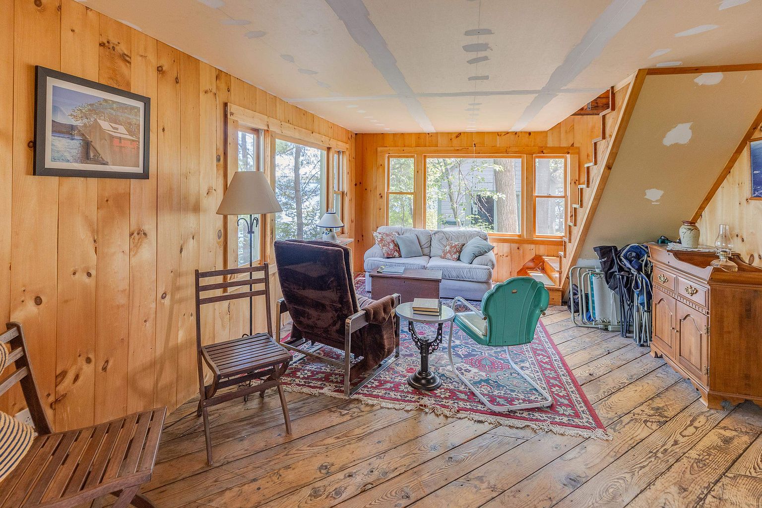 This rustic living room features warm wood-paneled walls and wide-plank wood flooring, creating a cozy, cabin-like atmosphere. The space is furnished with a mix of vintage-style seating, including a plush sofa, a dark upholstered armchair, and a retro teal chair, all centered around a patterned area rug. Large windows provide ample natural light and views of the surrounding trees, while a staircase tucked to the side adds architectural interest to the room.