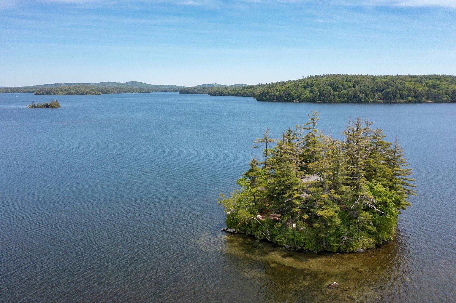 This aerial perspective captures a secluded island property nestled within a vast, calm lake. The island is densely forested with mature evergreen trees, partially concealing a private residence, and is surrounded by clear, shallow water that transitions into deep blue depths. The expansive landscape features rolling hills in the distance under a clear, bright sky, emphasizing the property's serene and private setting.