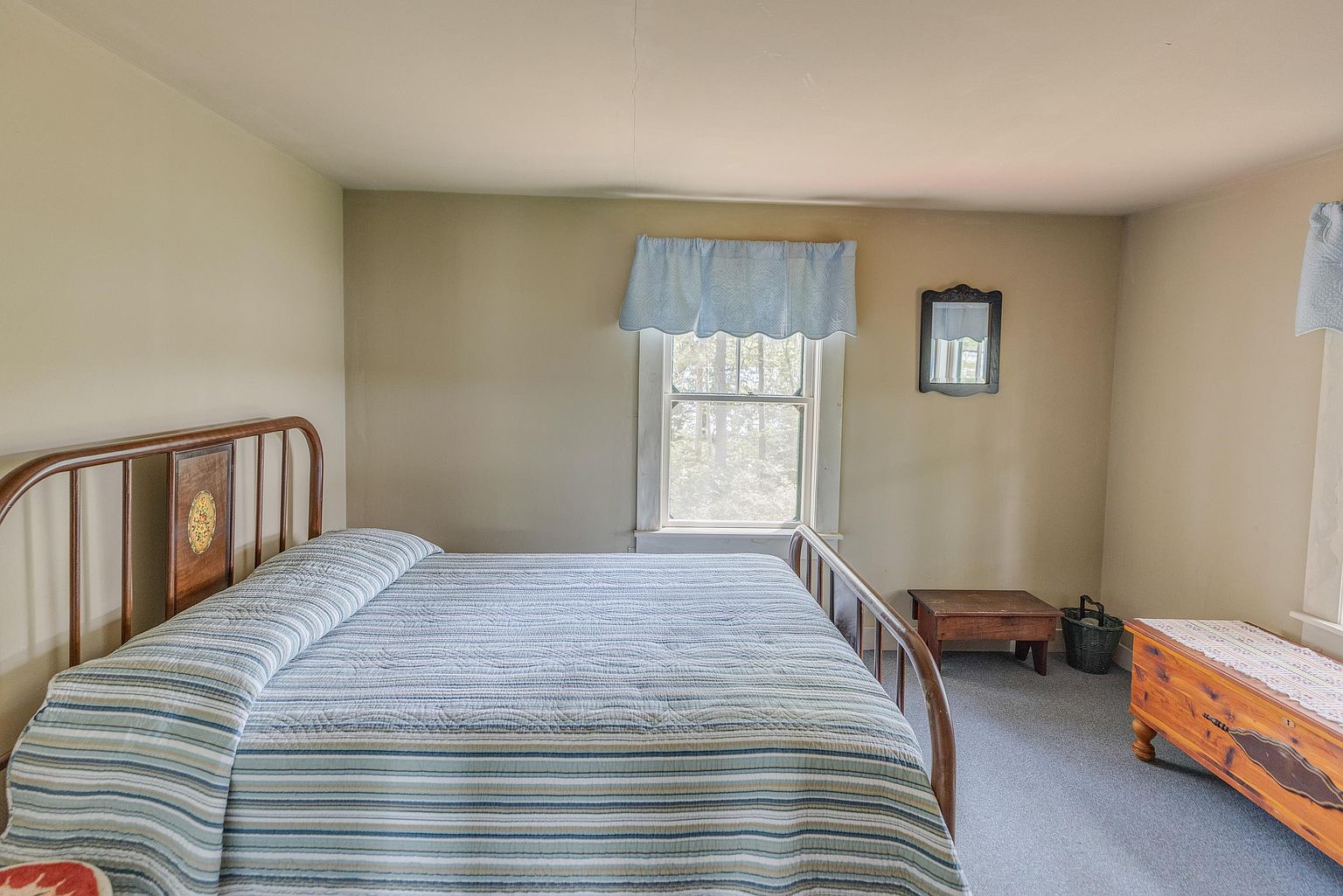This bedroom features a classic metal-framed bed dressed in a blue and white striped quilt, positioned centrally in the room. A window with a light blue valance provides natural light, while a wooden chest and a small side table offer storage. The room has a simple, traditional aesthetic with neutral wall tones and carpeted flooring.