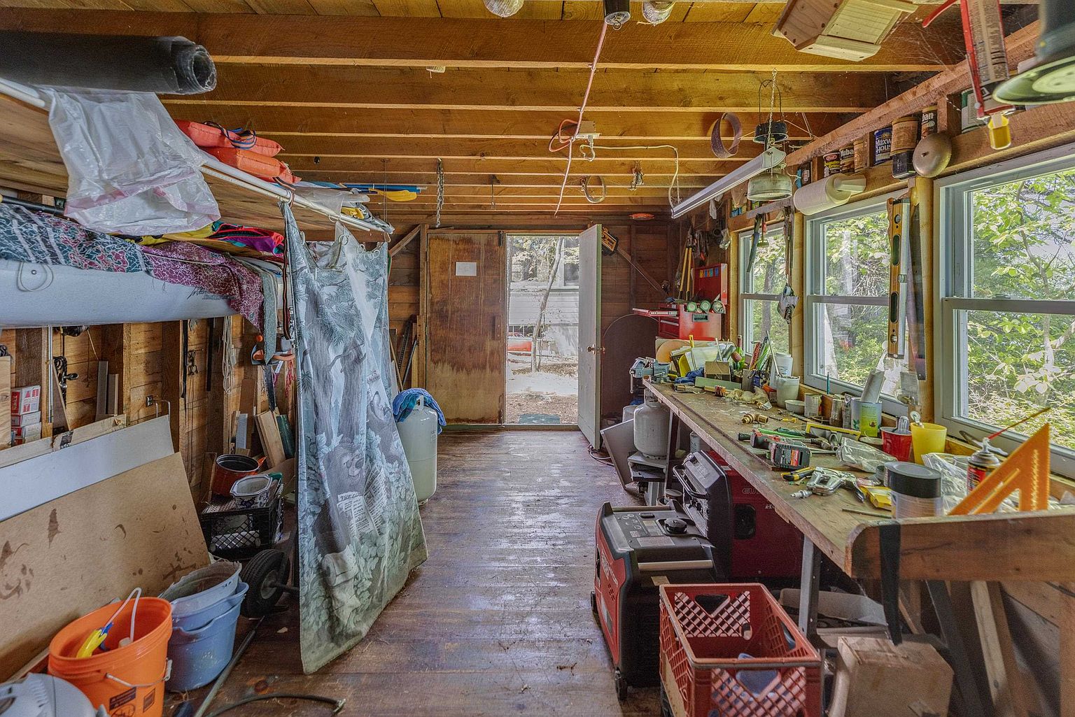 This rustic, wood-paneled workshop or garage space is filled with tools, equipment, and storage items, suggesting a functional hobbyist area. A long workbench runs along the right side under large windows that provide natural light, while an open doorway at the back reveals a view of the wooded outdoors. The space features exposed ceiling joists and a worn wooden floor, creating a utilitarian and industrious atmosphere.
