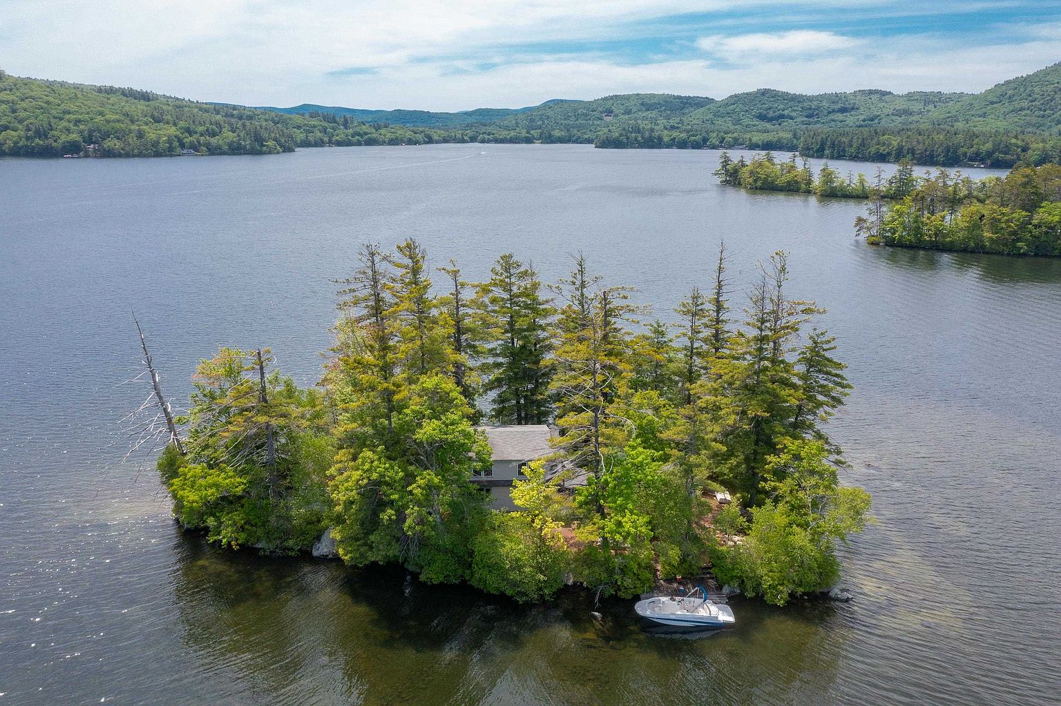 This aerial view captures a secluded private island retreat nestled in the middle of a large, calm lake. The property features a modest house surrounded by dense, mature trees, with a small boat docked at a wooden pier along the shoreline. The perspective provides a cinematic sense of isolation and tranquility, highlighting the natural beauty of the surrounding water and distant forested hills.