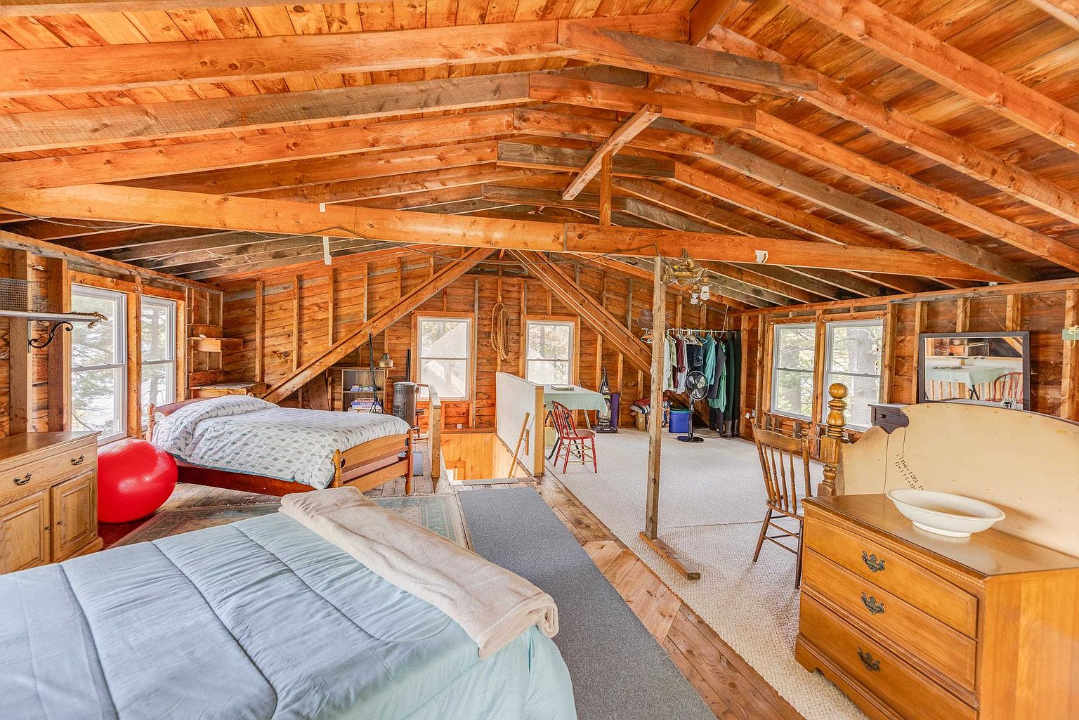 This spacious, rustic attic bedroom features exposed wooden rafters and beams, creating a warm and cozy atmosphere. The room is furnished with two beds, a wooden dresser, and a red exercise ball, with a carpeted area leading to a small seating space near the windows. The open-concept layout provides a clear view across the entire level, highlighting the natural wood construction and the charming, cabin-like aesthetic.