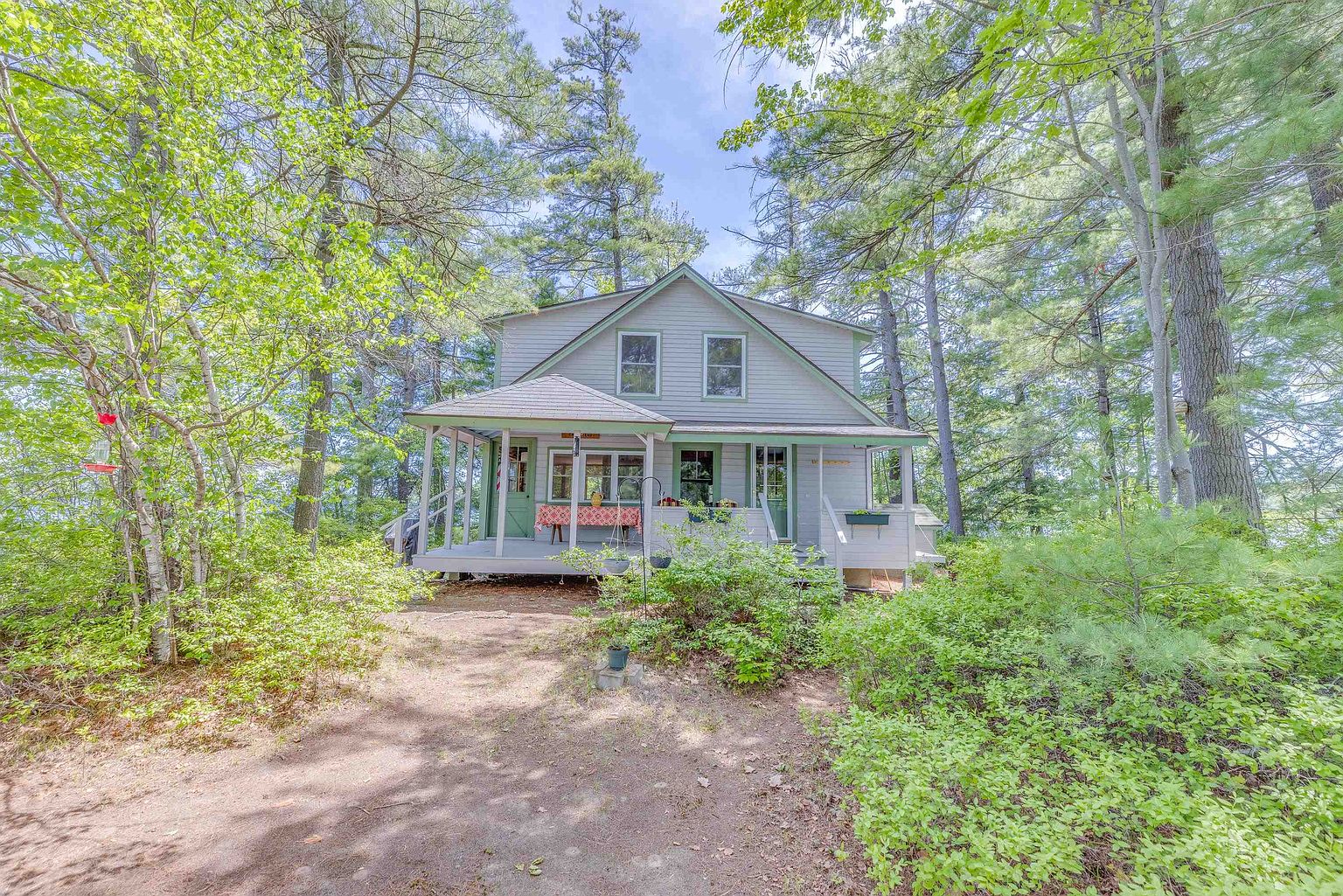 This charming, light-gray cottage is nestled within a lush, wooded setting, featuring a prominent covered front porch with a red-patterned tablecloth on a dining table. The home is framed by tall pine trees and dense greenery, creating a secluded and peaceful atmosphere. The perspective is a straight-on eye-level shot that highlights the home's rustic appeal and its integration into the natural landscape.