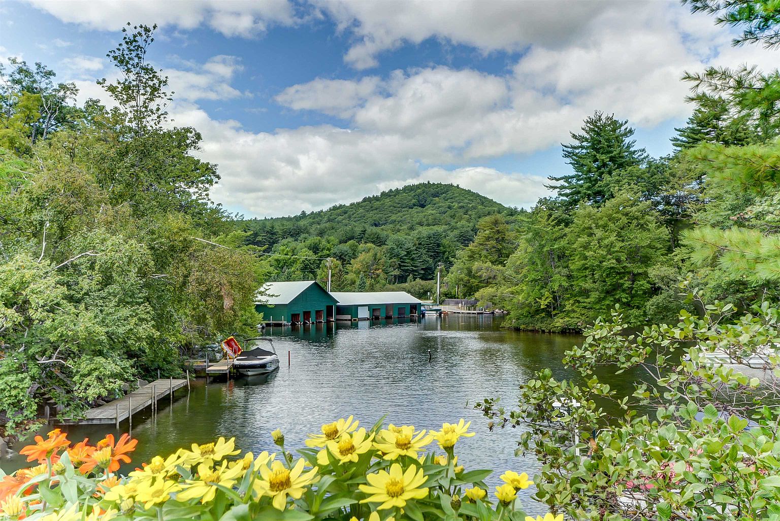 This scenic outdoor view captures a tranquil lakefront setting framed by vibrant yellow and orange flowers in the foreground. A long, green boathouse structure sits prominently on the water, surrounded by lush green trees and a gentle mountain slope in the background. The perspective is from a slightly elevated position, looking out over the water toward the boathouse, creating a peaceful and inviting atmosphere.