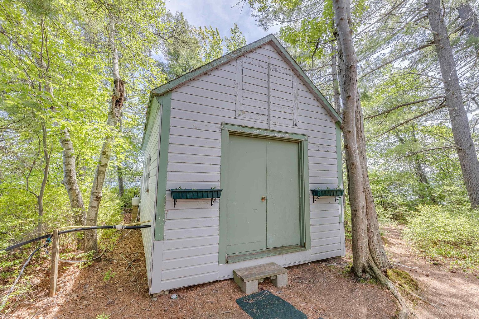 This image captures a small, light-colored wooden shed nestled in a wooded area, featuring double doors and two window boxes. The structure is set on a natural dirt path surrounded by mature trees, creating a rustic and secluded atmosphere. A small wooden step sits at the entrance, and a black mat is placed on the ground, emphasizing the shed's functional role in a natural landscape.