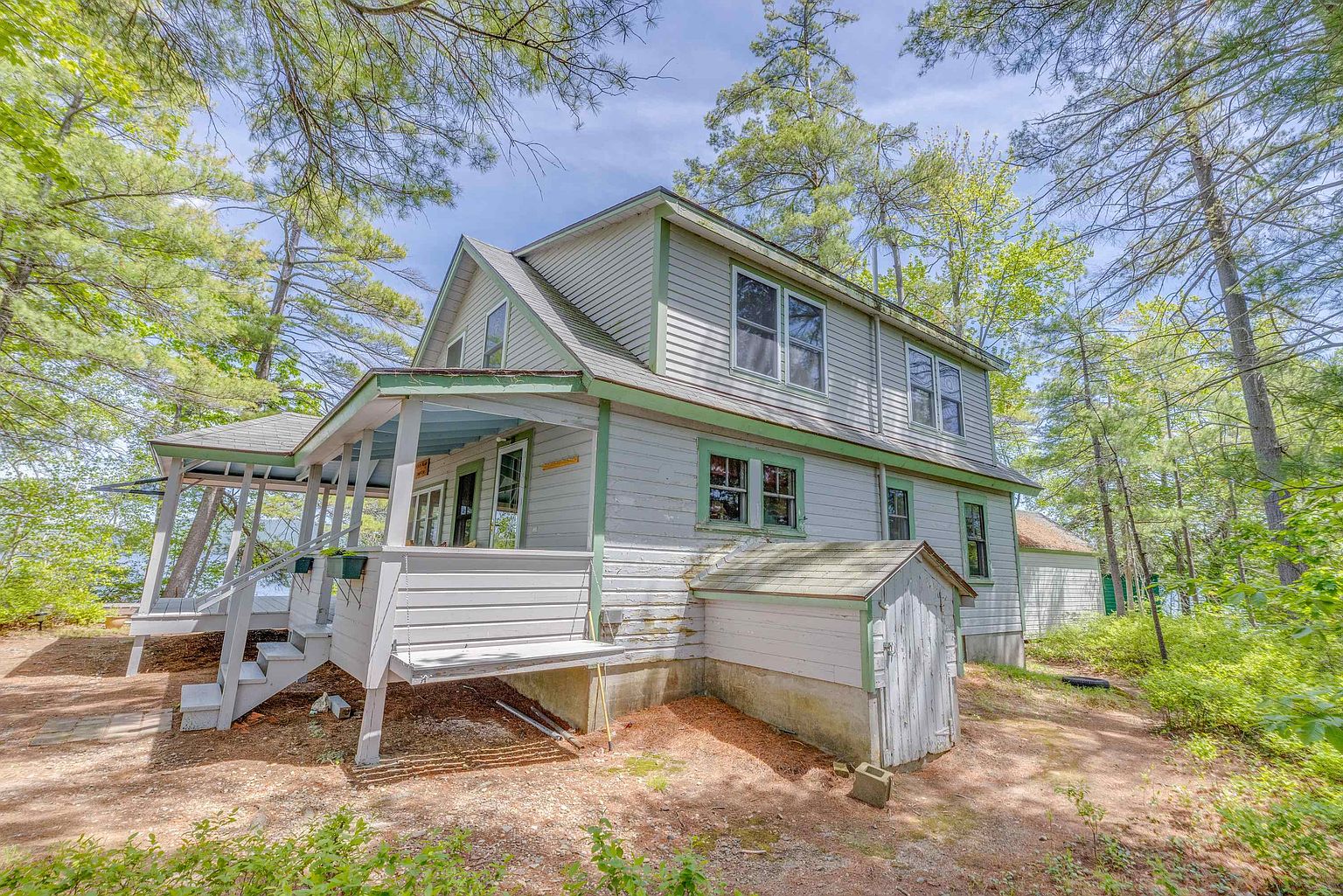 This side view of a rustic, light-gray wood-sided home captures a charming, multi-level structure nestled within a dense, wooded landscape. The exterior features a wrap-around porch with a screened-in section, a small shed-like structure attached to the side, and multiple windows that allow for natural light. The perspective is from ground level, emphasizing the home's integration into the natural, forest-like setting.