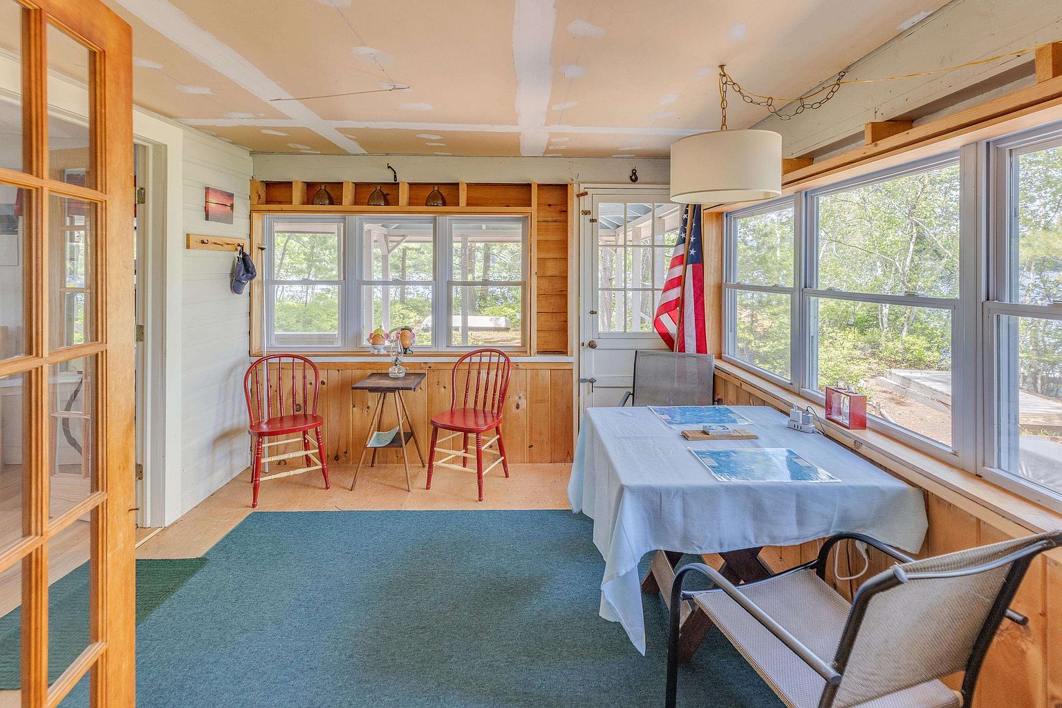This sunroom features a rustic, casual aesthetic with wood-paneled walls and a large, green area rug covering the floor. The space is furnished with a dining table covered by a light blue cloth and two red wooden chairs near a small side table, creating a cozy, informal seating area. Large windows line the walls, providing ample natural light and views of the surrounding trees, while a hanging pendant light fixture adds a touch of charm to the room.