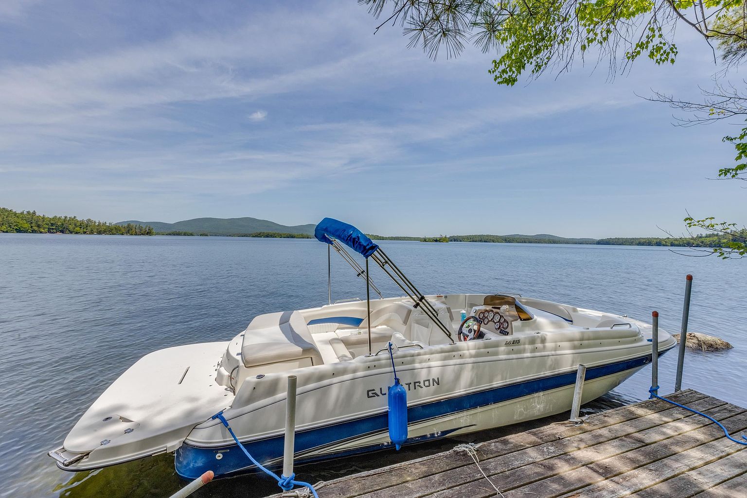 A white and blue Glastron DS 215 motorboat is docked at a wooden pier on a calm, expansive lake. The scene captures a serene waterfront setting with distant mountains under a bright, clear blue sky, emphasizing the recreational lifestyle associated with the property. The perspective is from the dock, looking out over the water toward the boat and the scenic horizon.