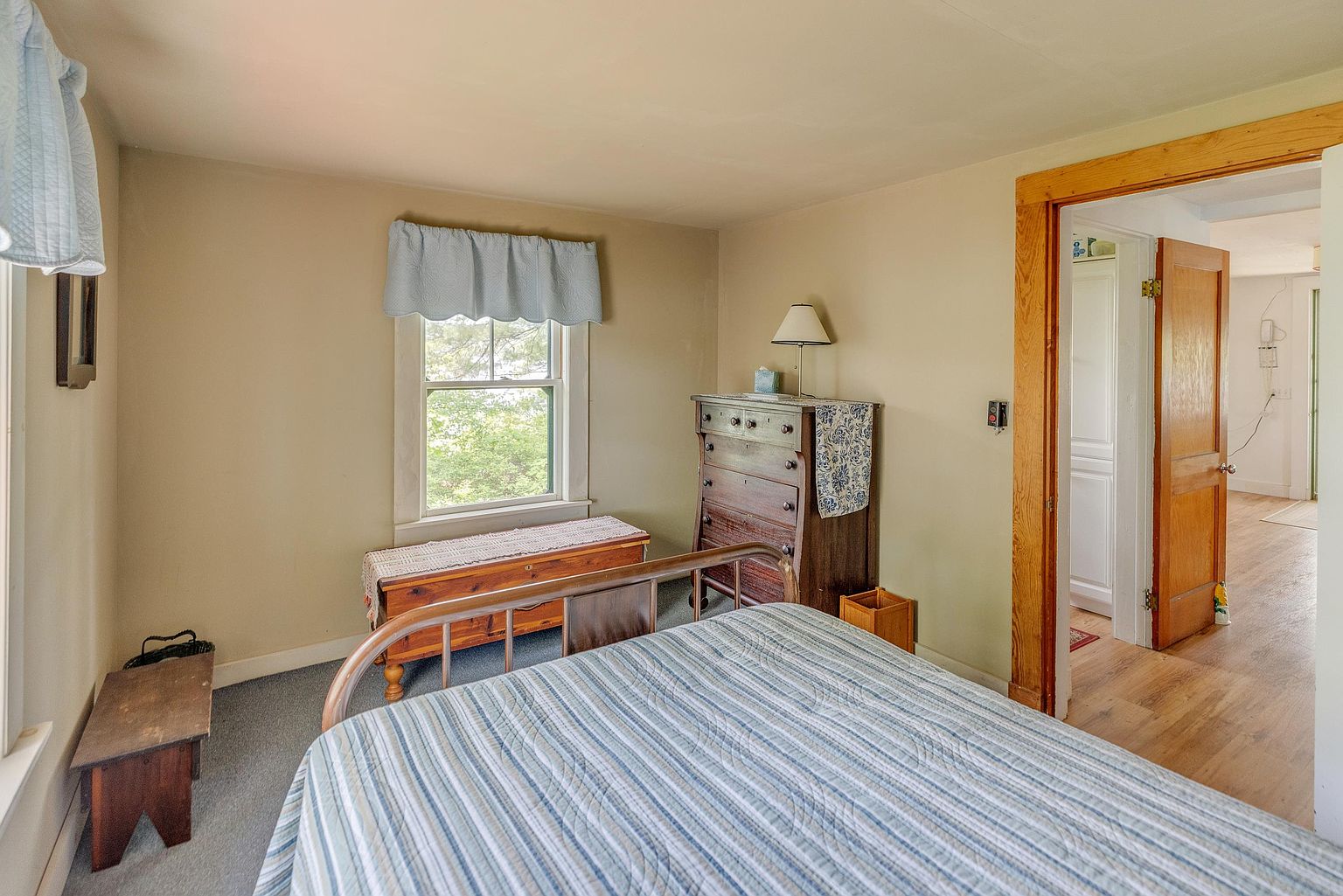 This cozy bedroom features a metal-framed bed with a blue and white striped comforter as the central focus. A wooden chest sits beneath a window dressed in a light blue valance, while a tall wooden dresser with a lamp occupies the corner. The room is finished with neutral-toned walls and carpet, leading into an adjacent room through a wooden doorway.