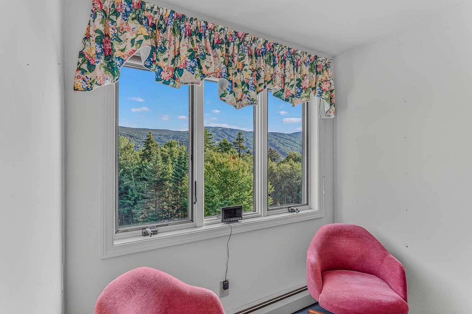 This bright room features a large triple-pane window that frames a scenic view of lush green mountains under a clear blue sky. Two vibrant pink armchairs are positioned in the foreground, creating a cozy seating area beneath a floral-patterned valance. The space is finished with simple white walls and a baseboard heater, offering a peaceful and inviting atmosphere.