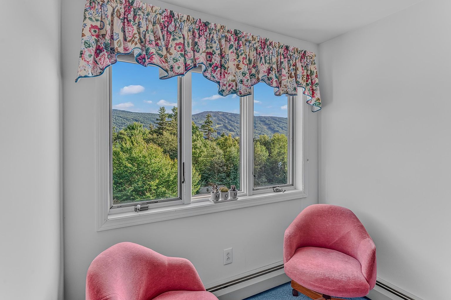 This cozy sitting area features two plush, pink velvet armchairs positioned in front of a large window that offers a scenic view of lush green mountains. The window is framed by a floral-patterned valance, and a small windowsill displays decorative 'MOM' lettering. The room is finished with neutral white walls and blue carpeting, creating a bright and inviting space.