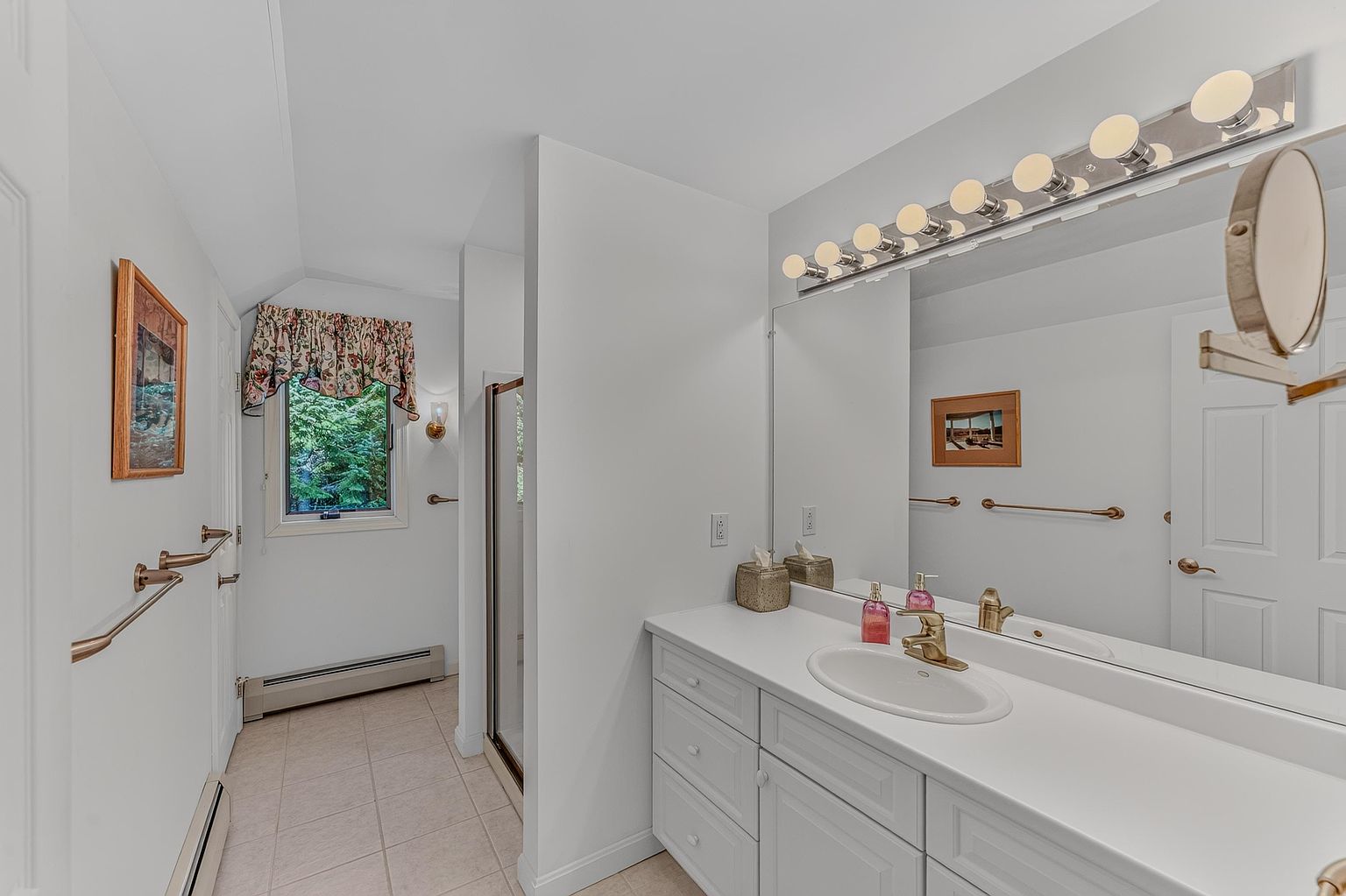 This bright bathroom features a white vanity with a single sink, gold-toned hardware, and a large wall-to-wall mirror illuminated by a multi-bulb vanity light. To the left, a tiled floor leads toward a shower enclosure and a window dressed with a floral valance, providing a view of the outdoors. The space is finished with neutral walls and gold-toned towel bars, creating a clean and functional aesthetic.