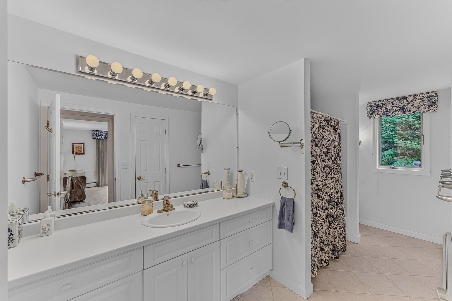 This bright and functional bathroom features a long white vanity with a single sink and a large, wall-to-wall mirror that enhances the sense of space. A floral-patterned shower curtain adds a decorative touch, while the neutral tile flooring and clean white walls create a cohesive, airy atmosphere. The room is well-lit by a multi-bulb vanity light fixture and benefits from natural light streaming through a window with a matching floral valance.