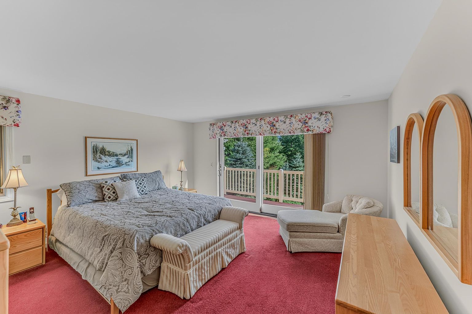 This spacious primary bedroom features a large bed with a patterned gray comforter, complemented by a matching upholstered bench at the foot. The room includes a light wood dresser with an arched mirror, a cozy chaise lounge near the sliding glass doors, and red carpeting throughout. Large sliding glass doors provide access to an outdoor deck and offer a view of the surrounding trees.