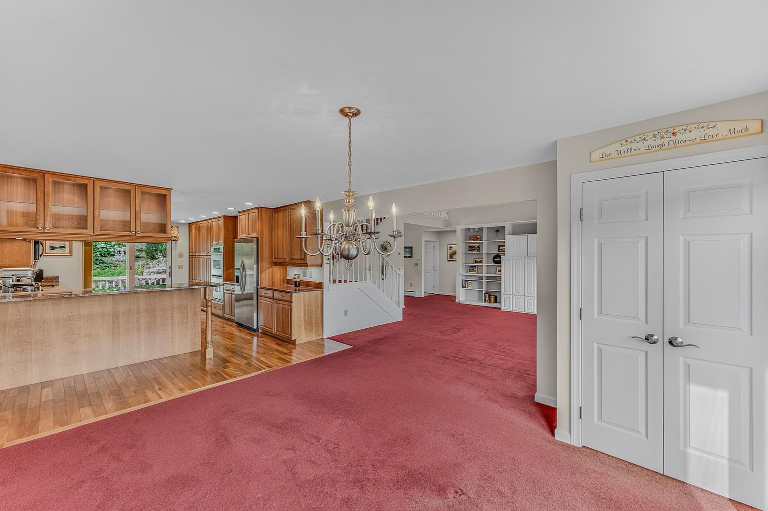 This open-concept dining area features a prominent chandelier as a central focal point, set against a backdrop of a kitchen with light wood cabinetry and a breakfast bar. The space transitions from a hardwood floor in the kitchen to a large carpeted area, leading toward a staircase and a built-in shelving unit in the background. A set of white double doors on the right adds a formal touch, while the neutral wall color creates a bright and airy atmosphere.