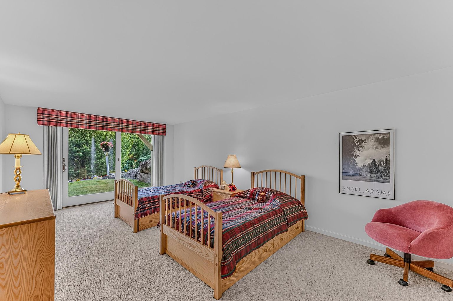 This guest bedroom features two twin beds with matching plaid bedding and light wood frames, positioned side-by-side. A large sliding glass door provides a view of the lush backyard, while a vibrant pink accent chair adds a pop of color to the neutral space. The room is completed by a framed Ansel Adams print on the wall and a light wood dresser, creating a cozy and inviting atmosphere.