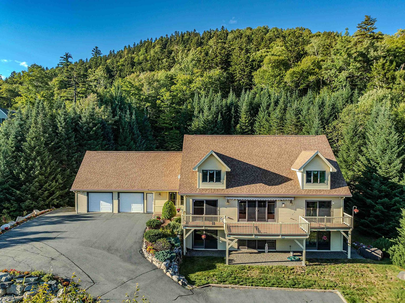This aerial perspective captures a two-story, light-yellow home nestled against a dense, lush forest backdrop. The property features a large asphalt driveway leading to an attached two-car garage, a spacious wooden deck spanning the rear, and a lower-level patio area. The home is characterized by its traditional gabled roofline, dormer windows, and a tiered landscape design with stone retaining walls.