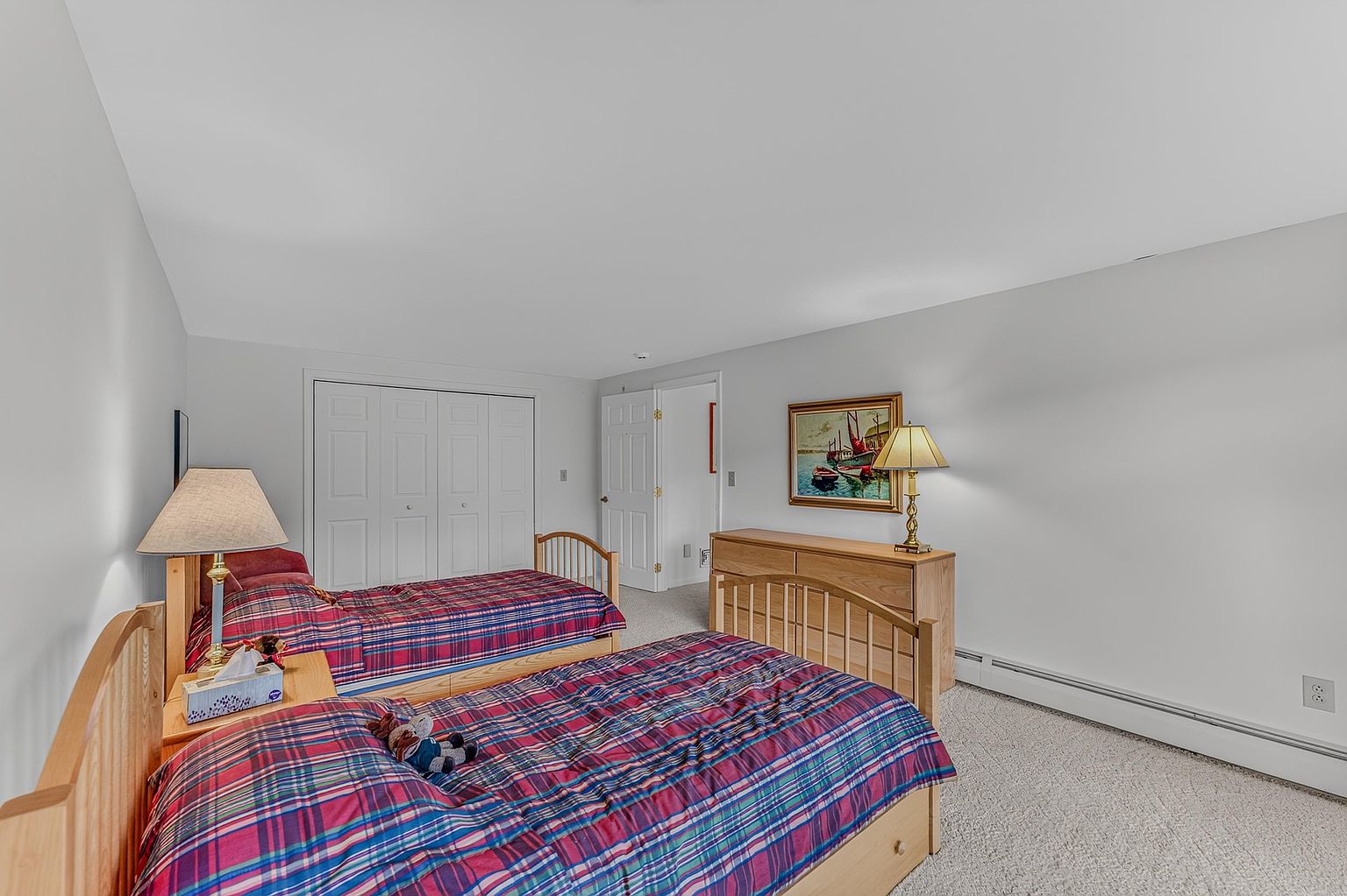 This guest bedroom features two twin beds with matching plaid bedding and light wood headboards. A wooden dresser sits against the wall, topped with a lamp and a framed painting, while a small nightstand with a lamp is positioned between the beds. The room is finished with neutral carpeting and white walls, creating a simple and functional space.