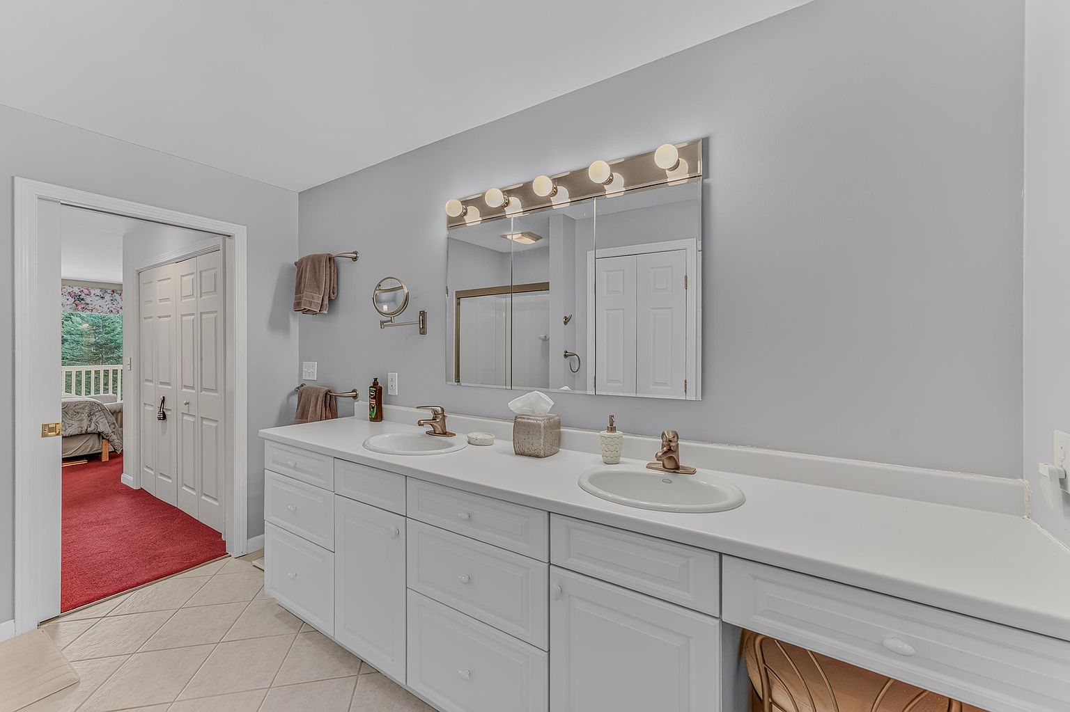 This primary bathroom features a long, white double-vanity with dual sinks and a large, multi-panel mirror illuminated by a five-bulb light fixture. The space is finished with light-toned walls and neutral tile flooring, creating a bright and functional atmosphere. A doorway on the left provides a glimpse into an adjacent bedroom with red carpeting, adding a touch of contrast to the otherwise neutral palette.