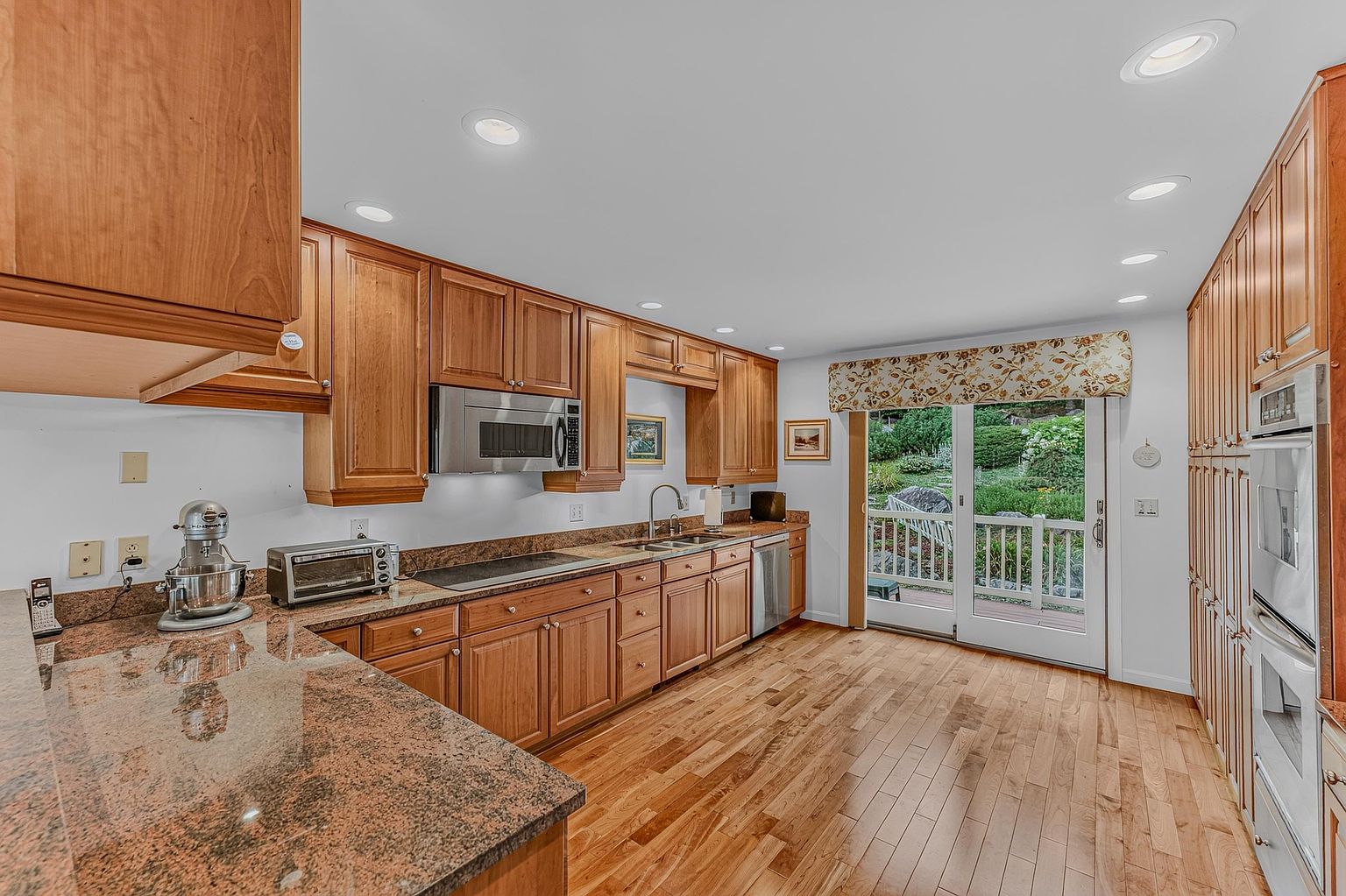 This well-lit kitchen features warm-toned wood cabinetry paired with speckled stone countertops and hardwood flooring. The space is equipped with stainless steel appliances, including a built-in oven and microwave, and offers a direct view of the lush backyard through a sliding glass door. The layout is functional and inviting, with recessed lighting throughout and a clean, traditional aesthetic.
