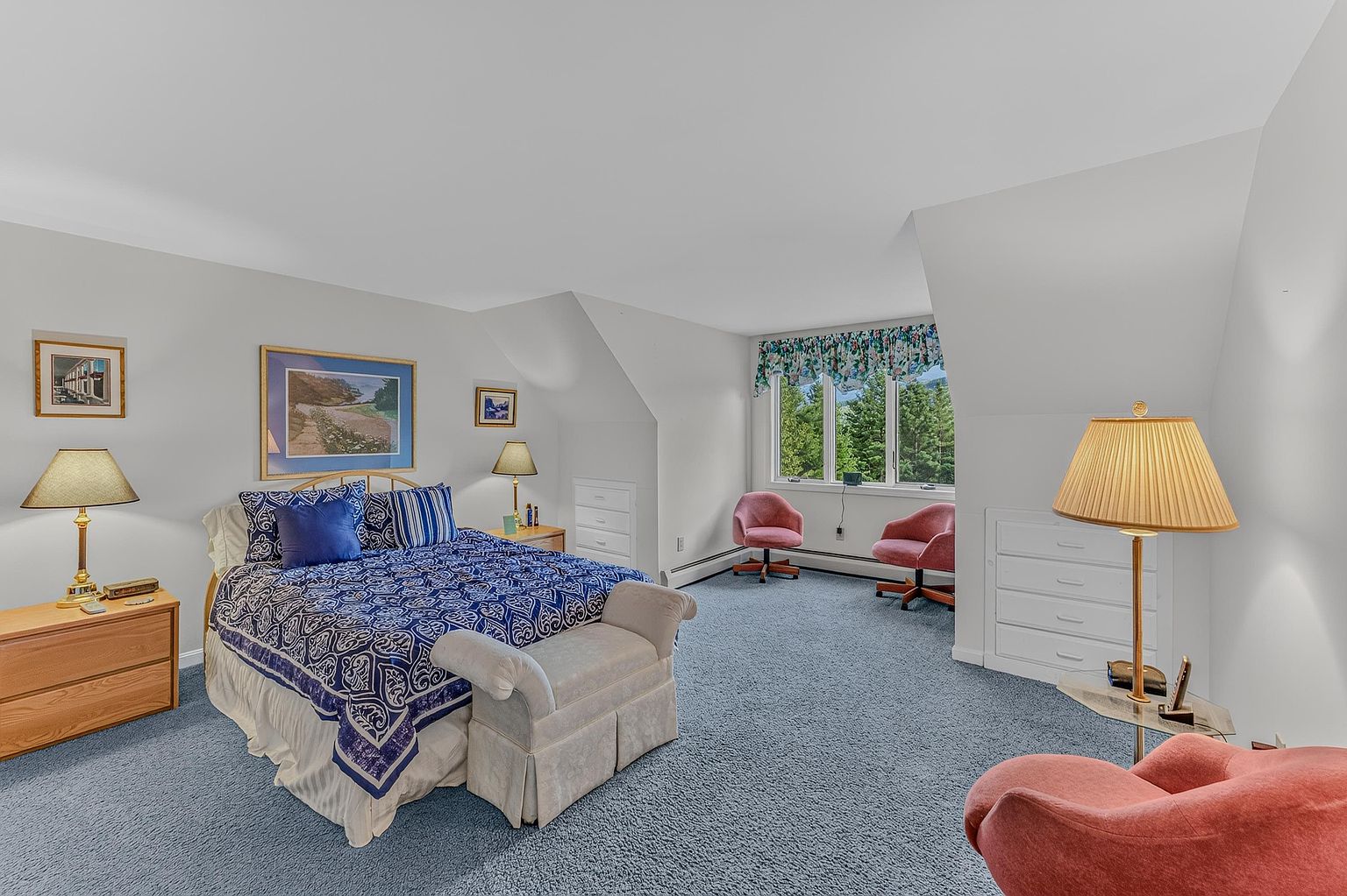 This spacious primary bedroom features a large bed with a blue and white patterned quilt, flanked by two light wood nightstands with matching lamps. The room includes a cozy seating area with two pink chairs near a window overlooking trees, and a third pink chair in the foreground. Built-in storage drawers are integrated into the walls under the sloped ceiling, creating a functional and organized living space.