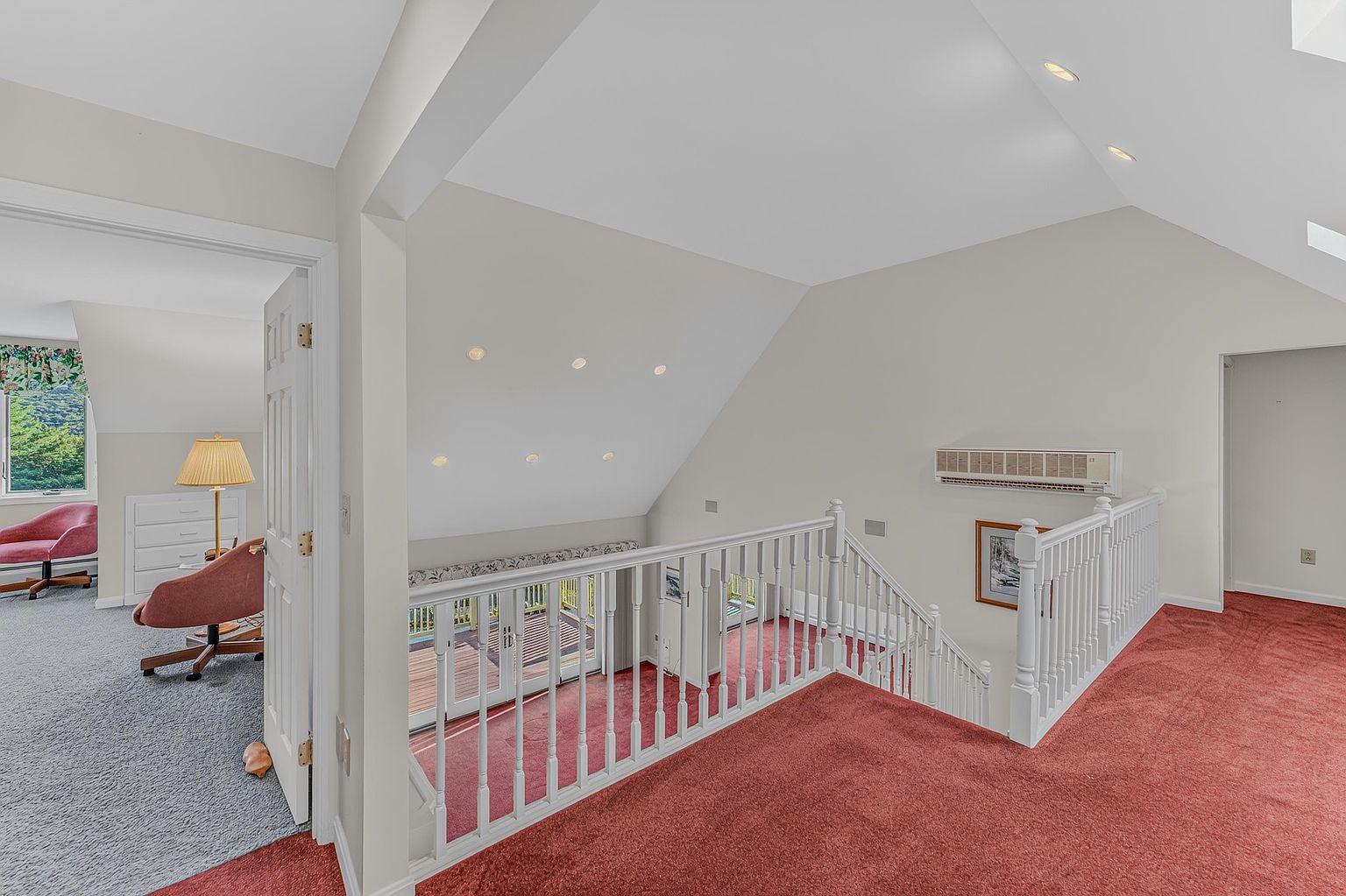 This interior view captures an open landing area featuring a red carpeted floor and a white wooden railing overlooking the level below. To the left, an open doorway leads into a carpeted bedroom with a pink armchair and a lamp, while a wall-mounted HVAC unit is visible on the right wall. The space is characterized by its vaulted ceiling with recessed lighting and a neutral color palette, creating an airy and transitional atmosphere.