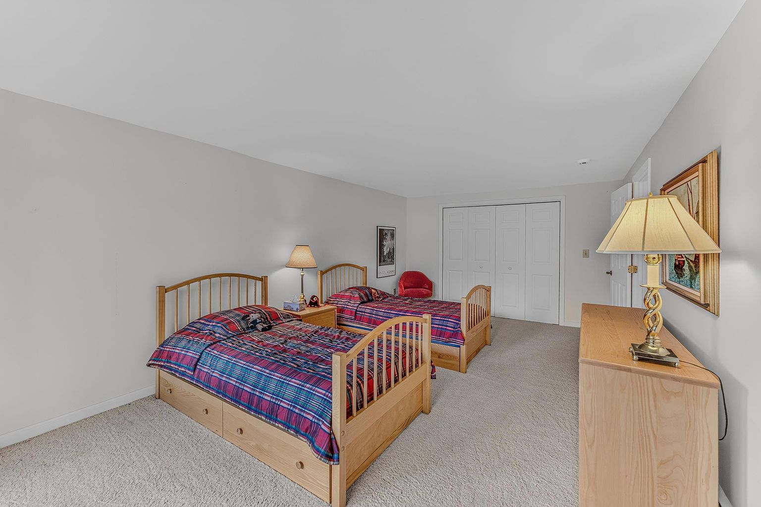 This spacious bedroom features two twin beds with light wood frames and plaid bedding, arranged side-by-side on neutral carpeting. A light wood dresser with a table lamp sits in the foreground, while a white closet door and a small red accent chair are visible in the background. The room is painted in a soft, neutral tone, creating a simple and functional living space.