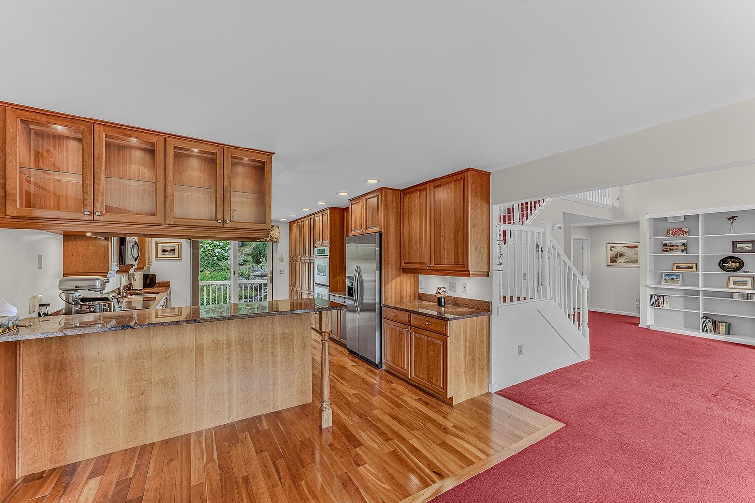 This kitchen features warm-toned wood cabinetry, granite countertops, and stainless steel appliances, including a refrigerator and wall oven. A peninsula with a breakfast bar provides additional workspace and seating, overlooking a living area with red carpeting and a built-in bookshelf. The open-concept layout connects the kitchen to the adjacent living space, creating a functional flow between the cooking and relaxation areas.